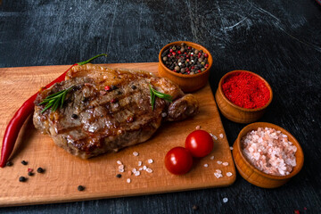 Grilled meat piece of ribeye beef meat on wooden cutting board on a wooden table stacked tomatoes garnished with hot peppers, peppercorns, paprika, himalayan salt. Fresh food. BBQ.