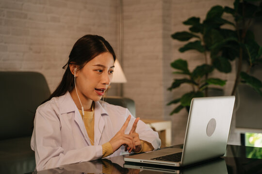 Successful Young Asian Female Doctor With Labcoat And Earphones Doing Video Conference Call For Consultation With Patient From Home At Late Night