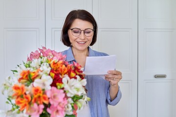 Happy middle aged woman with bouquet of flowers reading text from paper card