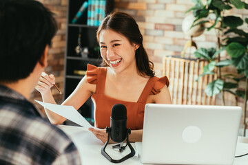 Cheerful young Asian female podcaster holding a questionnaire while interviewing male guest at workspace with mic and laptop