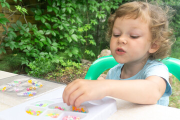 A little Caucasian girl, curly blonde, is captivated by creativity in the fresh air. The girl makes a homemade bead bracelet.