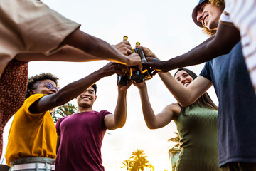 Happy multiracial friends having fun while chilling at sunset - Multiethnic young people toasting beer bottles on the beach - Celebration and summer party concept