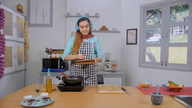 Happy young woman adding spices to the food in the pan while enjoying music. Wide shot of an Indian lady putting red chili powder  salt  and cumin powder to the dish in the frying pan