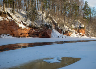 winter landscape with red sandstone cliffs on the bank of the river Salaca, the sun shines on the trees and the river bank, the ice covers the river, Latvia