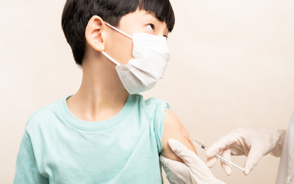 COVID 19 Vaccination For Kids And Children Concept. Cute Elementary Asian Boy With Face Mask Looking At Nurse While Receiving Vaccine Injection At Hospital. Herd Immunity, Safe, Protection.