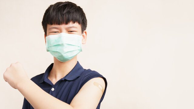 COVID 19 Vaccines for teenagers concept. Studio portrait of healthy Asian teenage boy with face mask, smile, fist up, show band aid on arm from first dose vaccine injection. Back to school campaign.