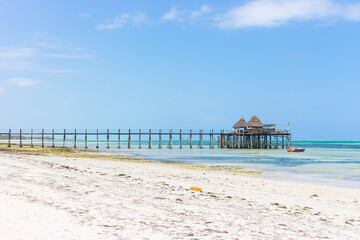 Wooden pier with boat and beach hut on sunny day. Low tide of Indian Ocean. Idyllic exotic resort. Pier in perspective with bungalows. Tropical paradise. Honeymoon in lagoon. Scenic tranquil seascape.