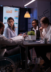 Overworked handicapped businessman in wheelchair sharing financial paperwork statistics overworking in business office meeting room. Diverse multi-ethnic brainstorming company ideas in evening