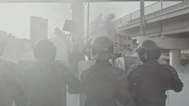Slowmo tracking shot of riot police with shields pushing back protesters with signs and smoke bombs