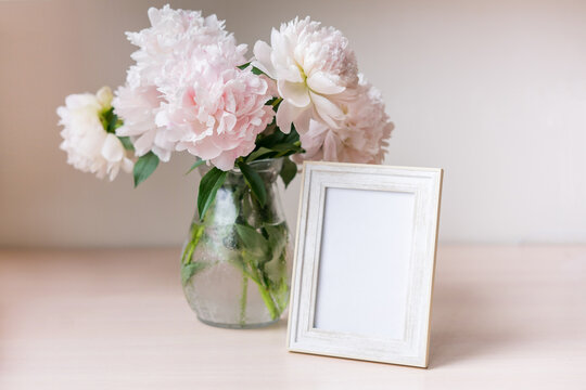 Portrait white picture frame mockup on wooden table. Modern glass vase with peony. White wall background. Scandinavian interior.	