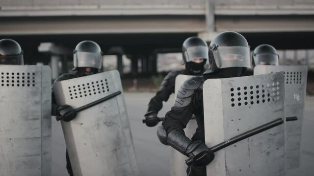 Slowmo shot of unrecognizable riot police officers in full gear beating their shields with batons while trying to intimidate protesters