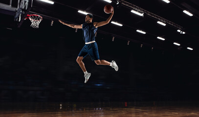 Young African sportsman, basketball player training in gym, idoors isolated on dark background. Concept of sport, game, competition. Slam dunk.
