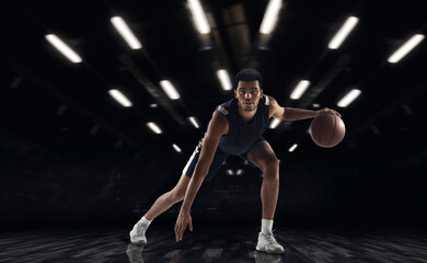 Fototapeta na wymiar Young African sportsman, basketball player training in gym, idoors isolated on dark background. Concept of sport, game, competition. Dynamic