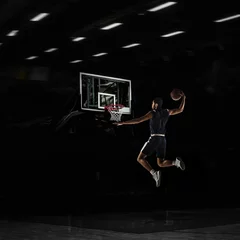 Fotobehang Young African sportsman, basketball player training in gym, idoors isolated on dark background. Concept of sport, game, competition. Slam dunk. © master1305