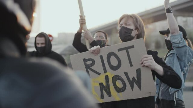 Handheld slowmo shot of angry masked people with signs chanting and confronting riot police outside