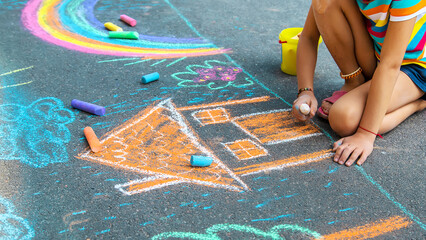 The child draws a house and a rainbow on the asphalt with chalk. Selective focus.