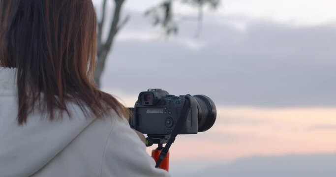 Rear view of woman photographer standing on the hill taking picture at the sunrise by using digital camera in slow motion shot. 