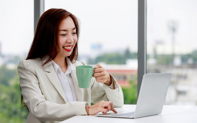Content young Asian female entrepreneur in stylish suit drinking coffee and using laptop while working on business project in modern workspace