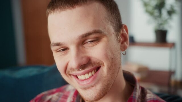 Portrait of brutal teenager with a nose piercing and earrings, brown-eyed happy man sitting on sofa and looking in camera smiling, British model posing. 
