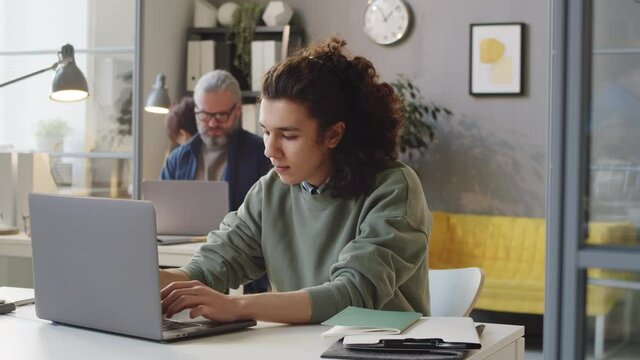 Tracking shot of young mixed race trainee in casualwear working on laptop at desk in office during probation period