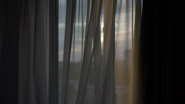 Light breeze blowing through opened window frame at sunset and transparent curtain swaying in dark room inside
