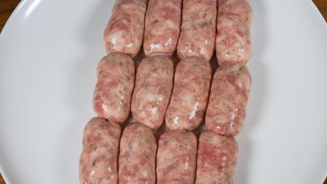 Close-up top view 4k stock video footage of many raw barbecue uncooked pork sausages with organic spices laying on big white plate standing on brown wooden table