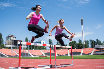 Two female sportswomen are jumping over an obstacle. Running with hurdles. Active lifestyle
