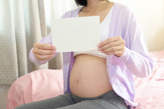 A pregnant Asian woman wearing casual clothes is sitting and show a blank white paper card in the bedroom. Image for presentation.