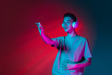Portrait of Caucasian young man in headphones isolated on dark red studio background in neon pink light. Concept of human emotions, facial expression.
