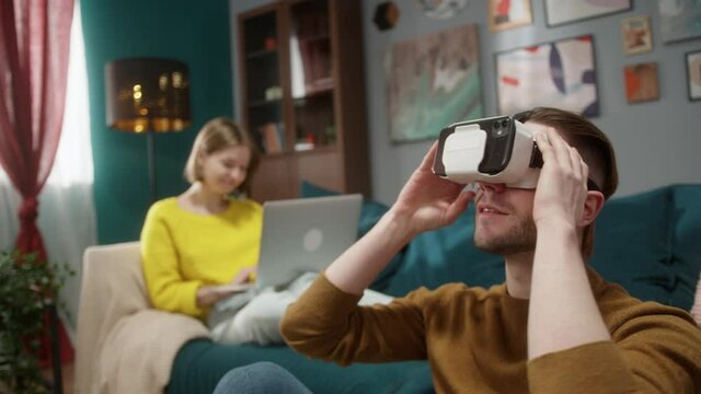 Young happy couple at home, man sitting on floor having fun playing VR headset video games and use technology virtual reality at home, woman on sofa with laptop share ar content to boyfriend