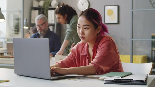 Young Asian female trainee with pink hair working on laptop at desk in office
