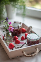 Jars with strawberry jam, strawberries and flowers on a linen towel and a white wooden tray.