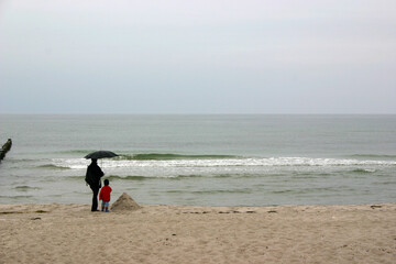 Eine Frau und ein Kind unter Regenschirm am Strand stehend blicken auf das Meer