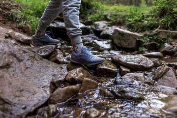 Close up of woman's shoes hiking in the mountains. Woman legs hiking at mountain wood. Outdoors crossing rock stones on river creek. Female travel on nature landscape.