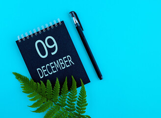 December 9th. Day 9 of month, Calendar date. Black notepad sheet, pen, fern twig, on a blue...
