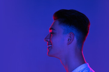 Close-up Caucasian young man posing isolated on blue studio background in neon pink light. Concept of human emotions, facial expression.