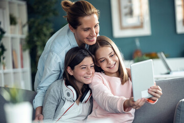 Beautiful mother and her little daughters making videocall with digital tablet while sitting together on couch at home.