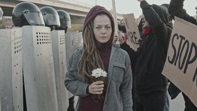 Portrait shot of young woman with dreadlocks holding flowers and looking at camera at protest People with signs chanting before unrecognizable riot police officers with shields
