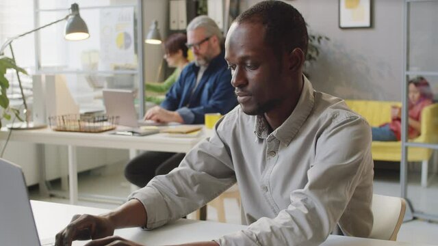 Portrait of Afro-American man in casualwear working on laptop and then posing for camera while sitting at desk in office