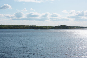 The opposite bank of a wide river with dense forest. Blue sky with clouds over the river.