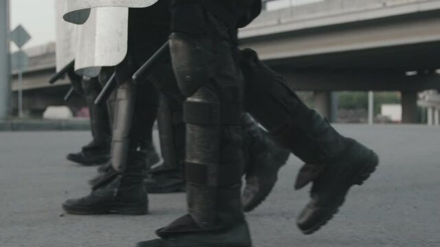 Low-section Tracking With Slowmo Of Unrecognizable Riot Police Officers In Gear Carrying Shields And Batons And Walking In Formation