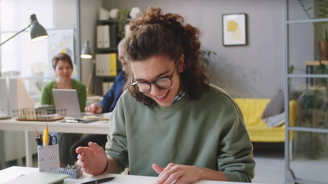 Young cheerful male trainee looking at camera, speaking and showing document while sitting at office desk and having business meeting on online video call