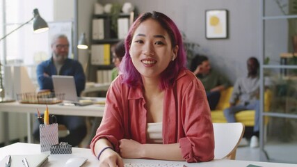Young beautiful Asian woman in wireless earphones sitting at office desk, looking and camera and speaking via online web call - Powered by Adobe