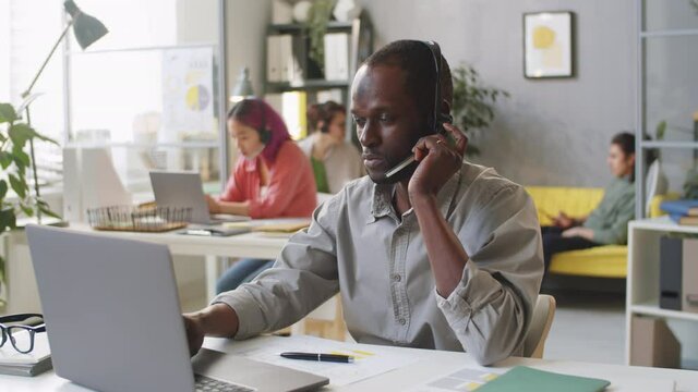 Young afro-american man in headset using laptop at desk and answering web call while working in office of support center