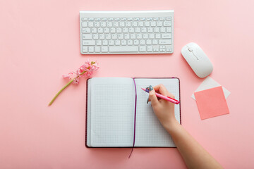 Pink work desk with female hand writes notes in notebook at workplace. Desktop office space top...