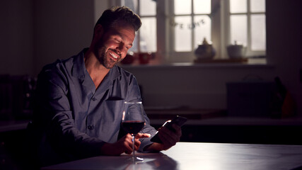 Man Wearing Pyjamas Sitting In Kitchen With Glass Of Wine At Night Using Mobile Phone