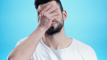 frustrated man obscuring face with hand isolated on blue