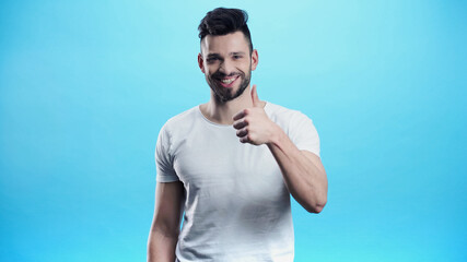 Naklejka premium smiling man looking at camera and showing thumb up isolated on blue