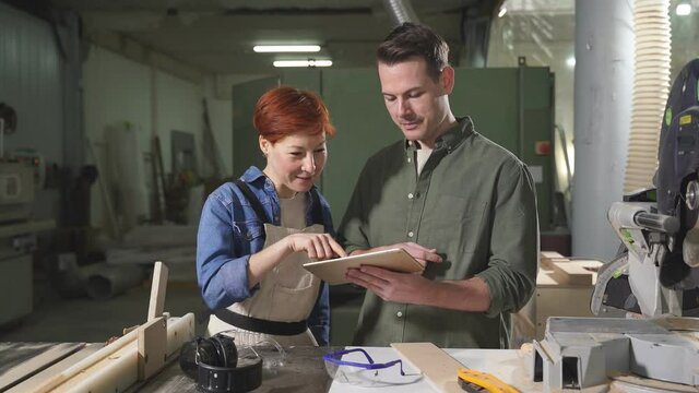 Two nice joiners discuss customer's orders, looking at screen of digital tablet, sharing opinions, smile.