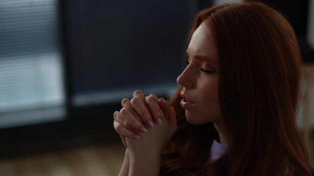 Close-up of young woman praying with eyes closed holding folded hands in front of face at home in dark living room. Tracking shot of worried female put hands in prayer praying excitedly with hope.